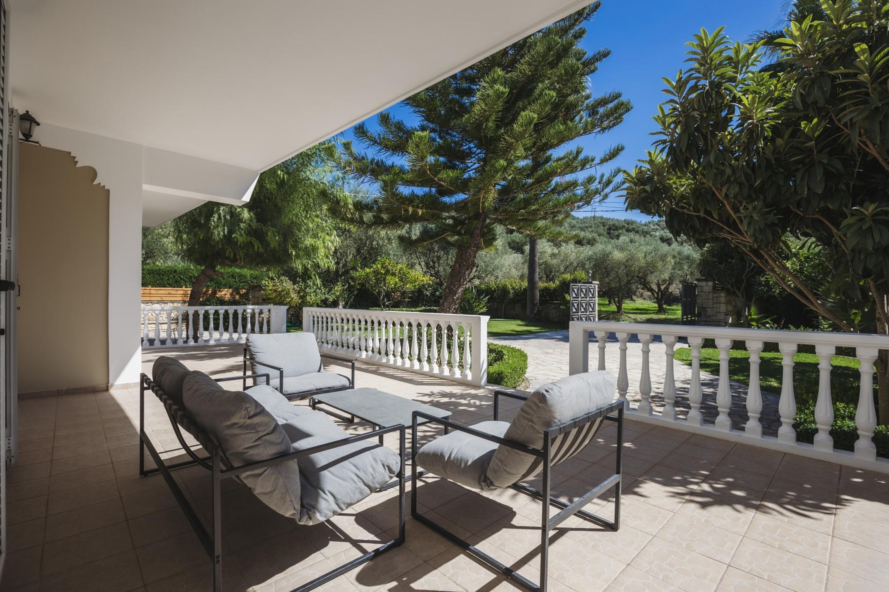 A covered outdoor seating area with a white railing, a pergola, and a view of a landscaped garden with trees and a pathway.