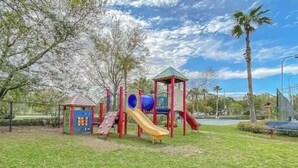 A playground with slides and climbing structures, surrounded by grass and trees.