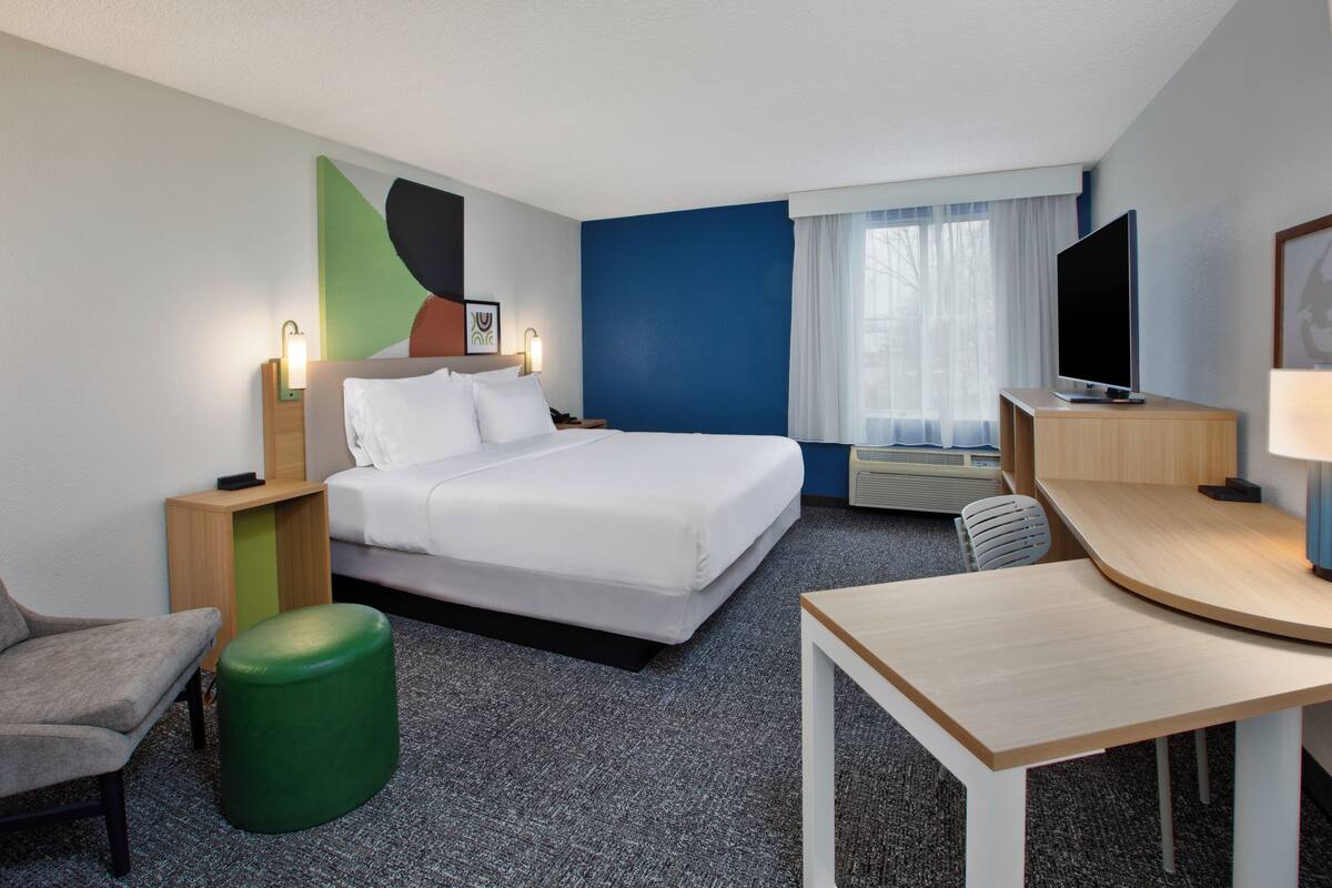 A hotel room with a bed, a desk, a chair, a green ottoman, a TV, and a window with curtains.