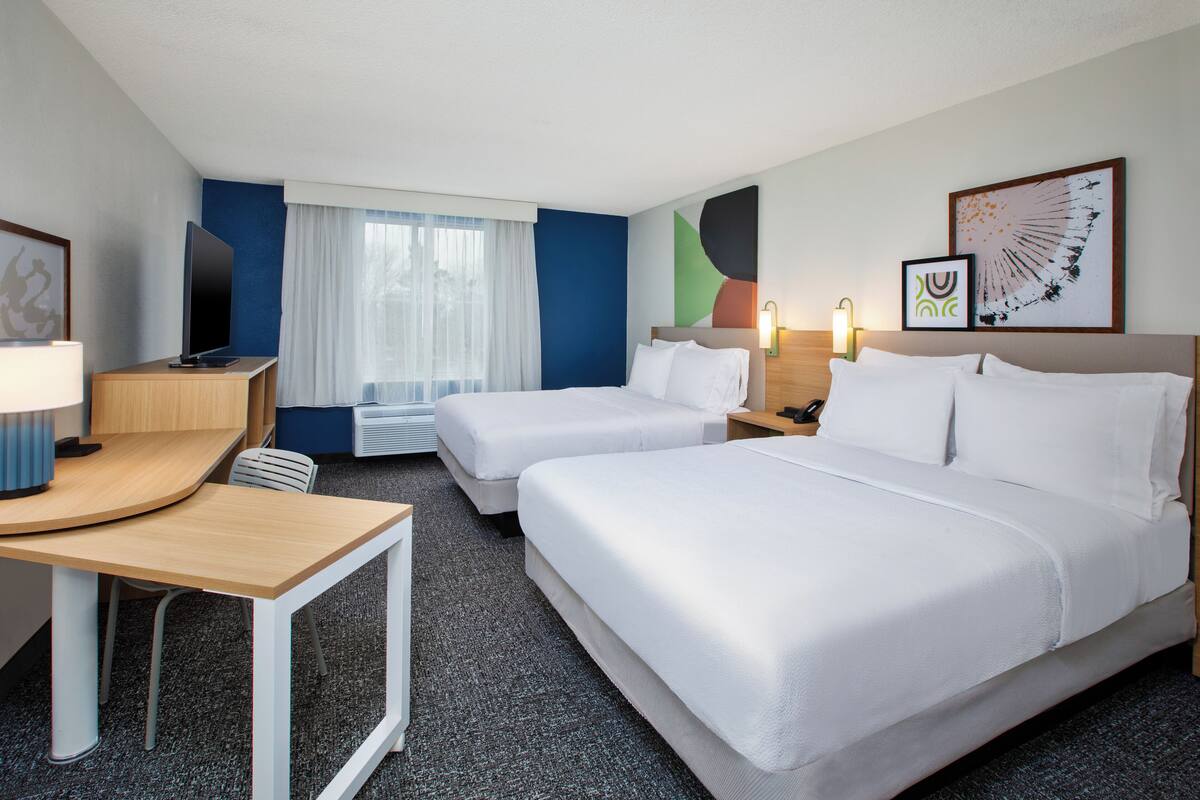 A hotel room with two beds, a desk, a TV, and a window with curtains.