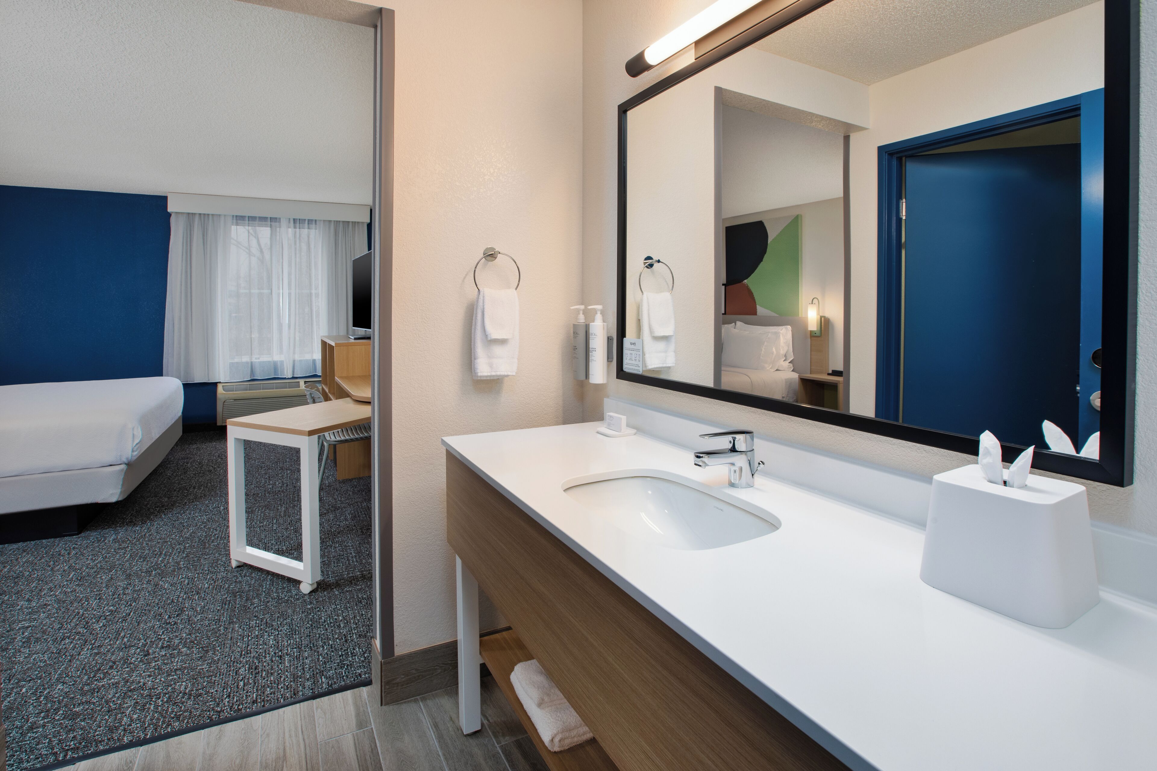 A hotel room with a bed, a bathroom with a sink and mirror, and a small table.