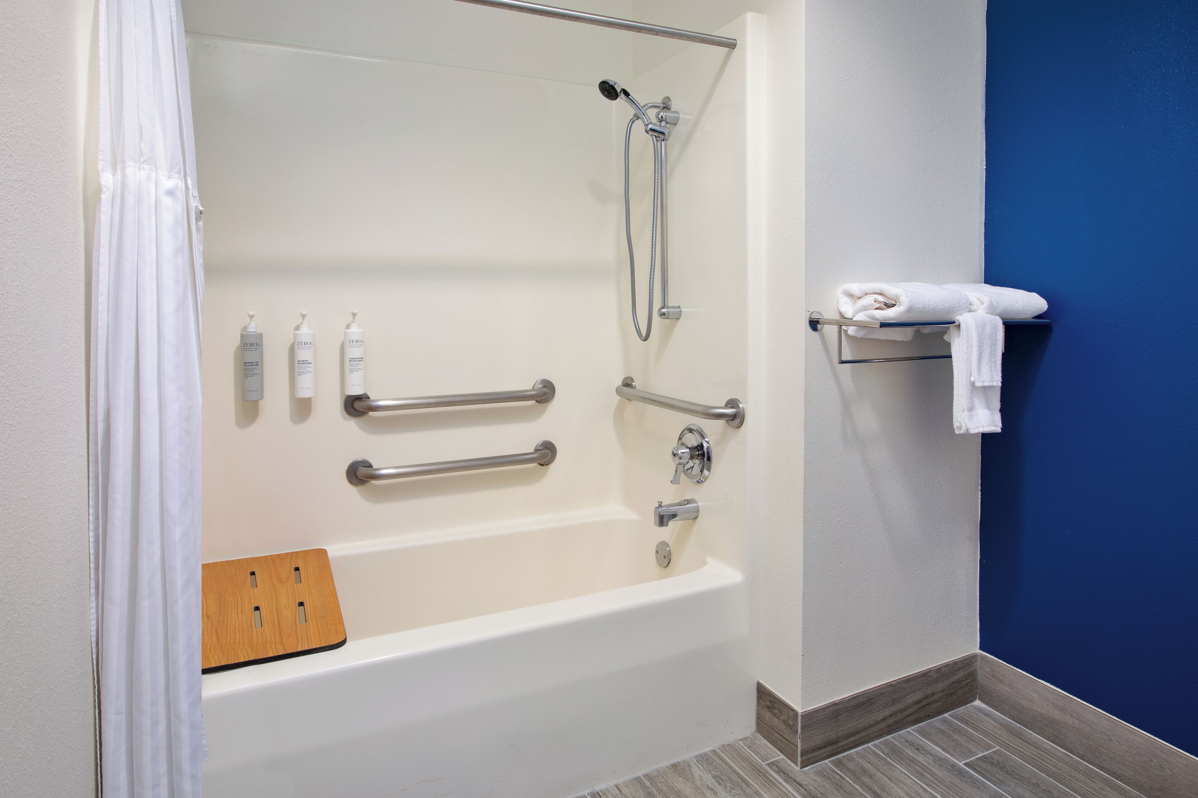 A modern bathroom with a walk-in shower, grab bars, a wooden bath mat, and a towel rack.