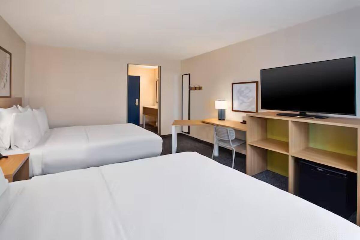 A hotel room with two beds, a desk, a television, and a small kitchenette.