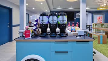 Una estación de café con tres dispensadores etiquetados 'Agua Caliente', 'Café Descafeinado' y 'Café Regular'. También hay condimentos y un letrero que dice 'Fogg's'.