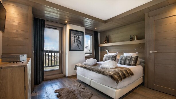 A modern bedroom with a large bed, wooden flooring, a fur rug, and a balcony with a city view. - Wifi, 150m from ski slopes, 2nd floor, view mountain, sauna, balcony, fireplace or stove, parking (Courchevel)