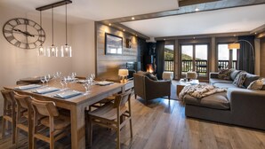 A spacious living area with a wooden dining table set for a meal, a comfortable sofa, and a fireplace. - Wifi, 150m from ski slopes, 2nd floor, view mountain, sauna, balcony, fireplace or stove, parking (Courchevel)