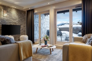 A cozy living room with a stone wall, a flat-screen TV, and a view of a snowy landscape through large glass doors.