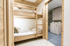 A wooden bunk bed with a ladder, a small bedside table, and a pillow. There is a kitchen area with cabinets and a countertop visible in the background.