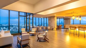 Un salón moderno con grandes ventanales que ofrecen vistas a la ciudad, piso de madera y mobiliario contemporáneo.