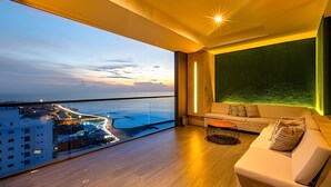 Un salón moderno con balcón que ofrece vistas a una ciudad costera al atardecer.