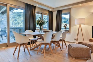 Ein stilvoller Essbereich mit Holztisch und weißen Stühlen, großen Fenstern mit Vorhängen und Blick auf eine gebirgige Landschaft.