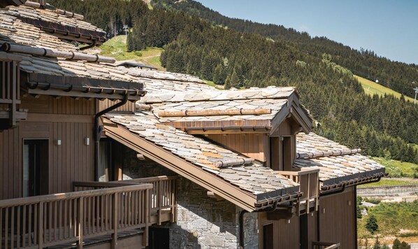 A wooden chalet with a sloped roof, stone foundation, and a balcony overlooking a mountainous landscape.