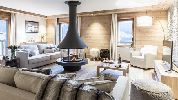 A cozy living room with a central fireplace, wooden walls, and a sofa with fur throw. - Wifi, 150m from ski slopes, 2nd floor, view mountain, sauna, balcony, fireplace or stove, parking (Courchevel)