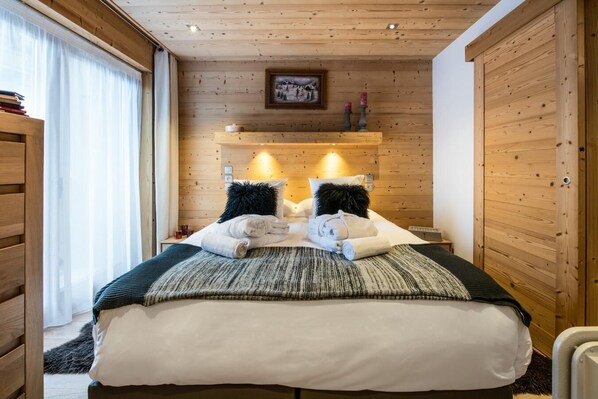 A wooden-paneled room with a bed, two wall-mounted lamps, a framed picture, and a window with curtains.