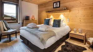 A modern bedroom with a wooden ceiling, a large bed with white linens, a chair, a nightstand with a lamp, and a view of the outdoors. - Wifi, 150m from ski slopes, 2nd floor, view mountain, sauna, balcony, fireplace or stove, parking (Courchevel)
