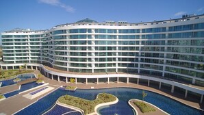 Un hôtel moderne à la façade incurvée, doté de nombreux balcons et d’un grand espace central avec piscine, entouré de plus petites piscines.