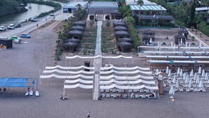 Vue aérienne d’un complexe hôtelier de bord de mer, avec des constructions à toits de chaume, des chaises longues et une zone de baignade.
