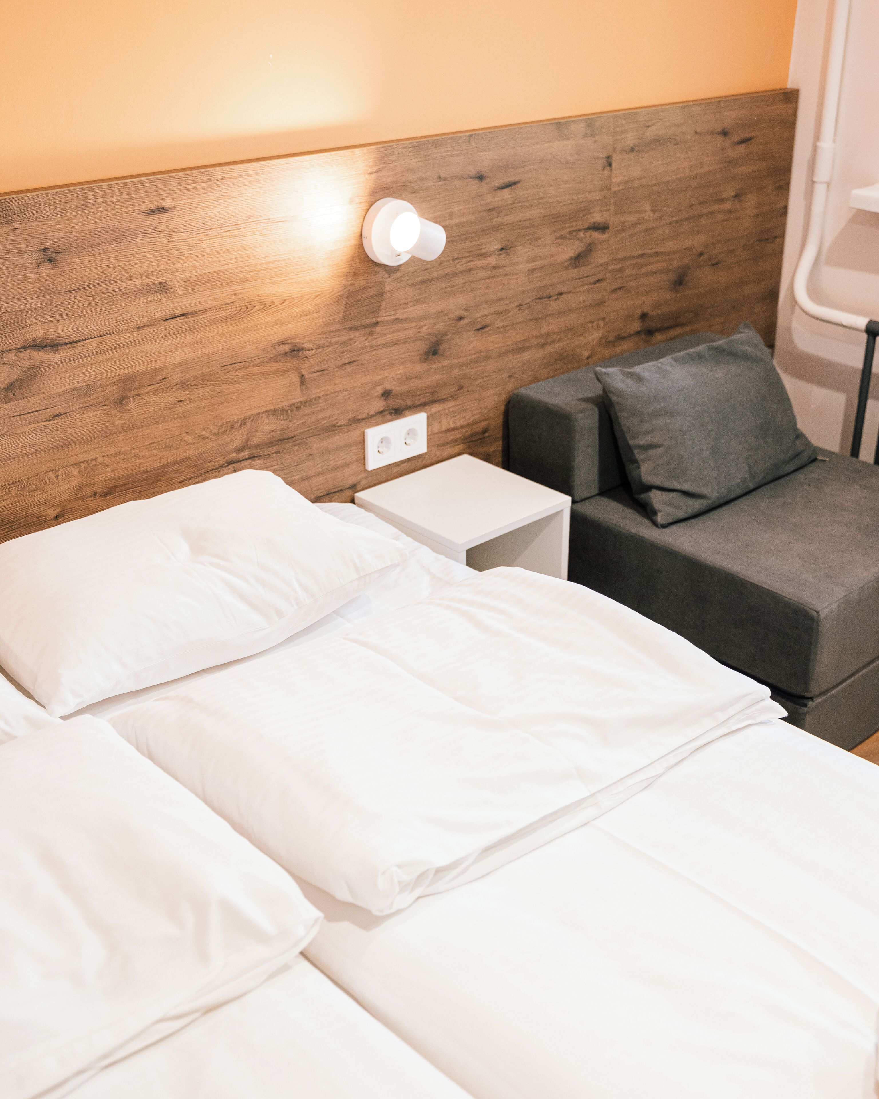 Una cama bien hecha con sábanas blancas, cabecera de madera, mesita de noche y sofá gris con un cojín.