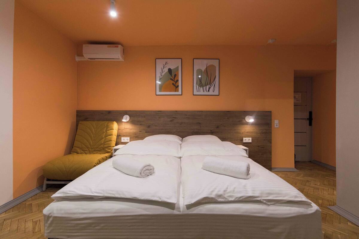 A double bed with white linens, a wooden headboard, two wall-mounted lights, and two framed artworks above the bed.