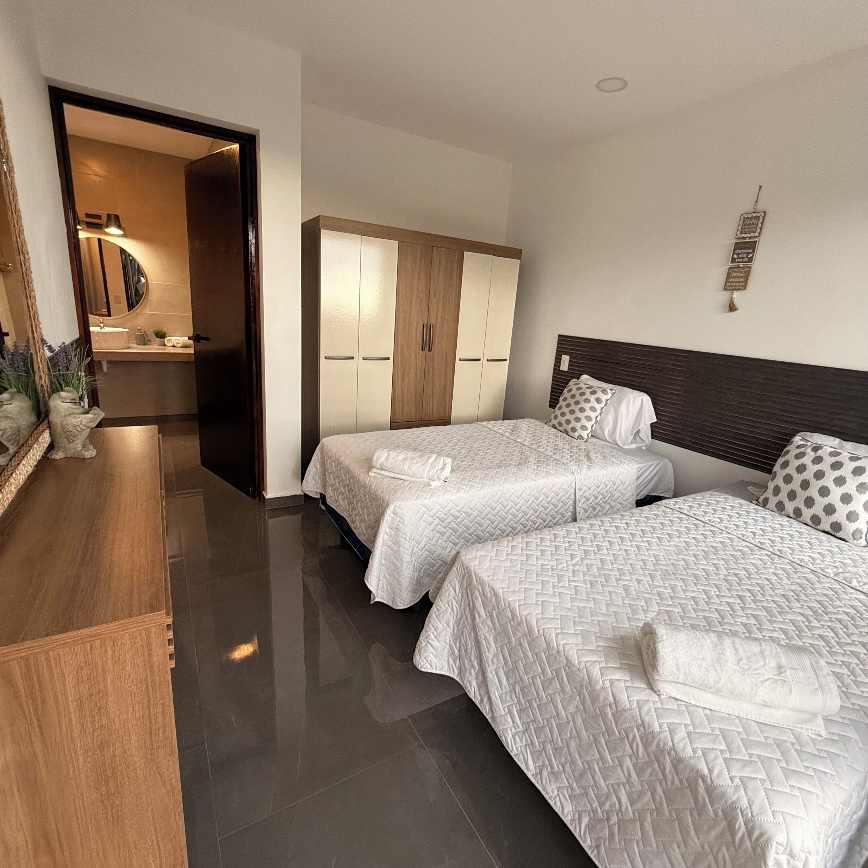 A hotel room with two beds, a wooden wardrobe, a mirror, and a bathroom with a sink and mirror.