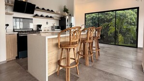 Una cocina moderna con una isla central, taburetes de bar, un televisor de pantalla plana y un amplio ventanal con vistas al verdor.