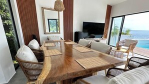 Un comedor moderno con una gran mesa de madera, sillas de mimbre y un amplio ventanal con vista al océano.