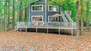 A two-story cabin with a deck, surrounded by trees and fallen leaves.