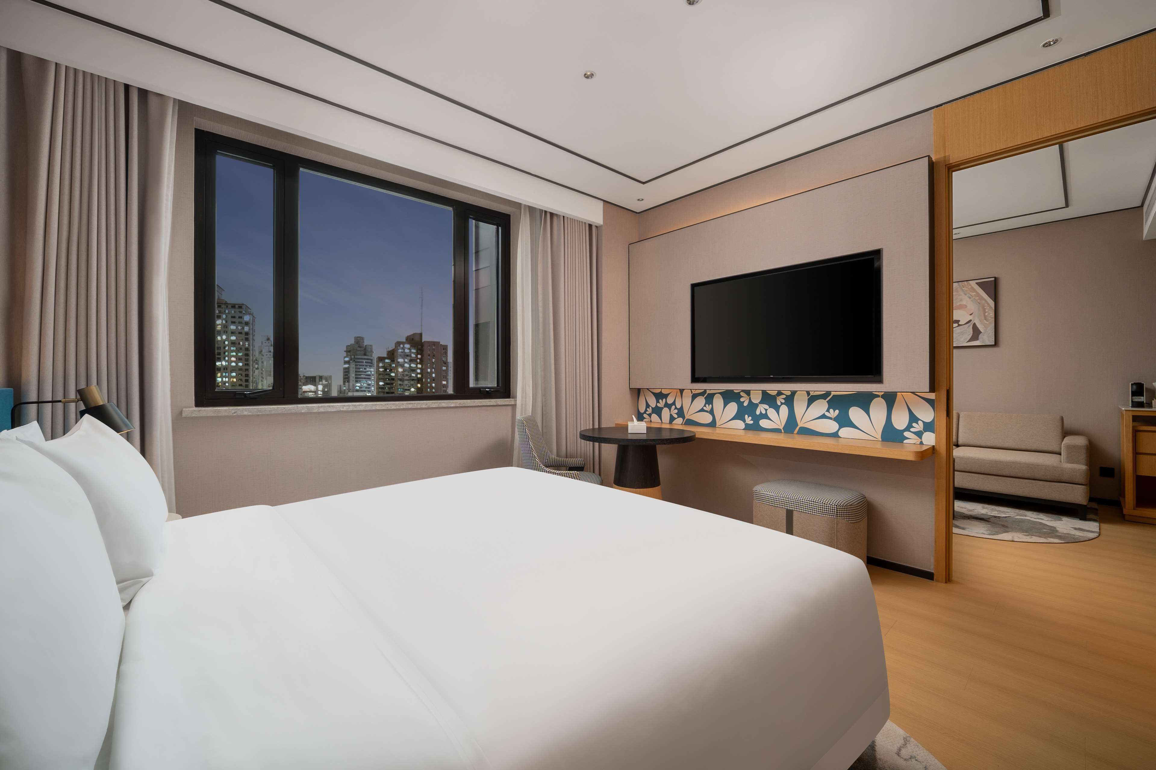 Une chambre d’hôtel avec un grand lit, un téléviseur fixé au mur, une petite table et une vue sur le panorama urbain, visible à travers une grande fenêtre.