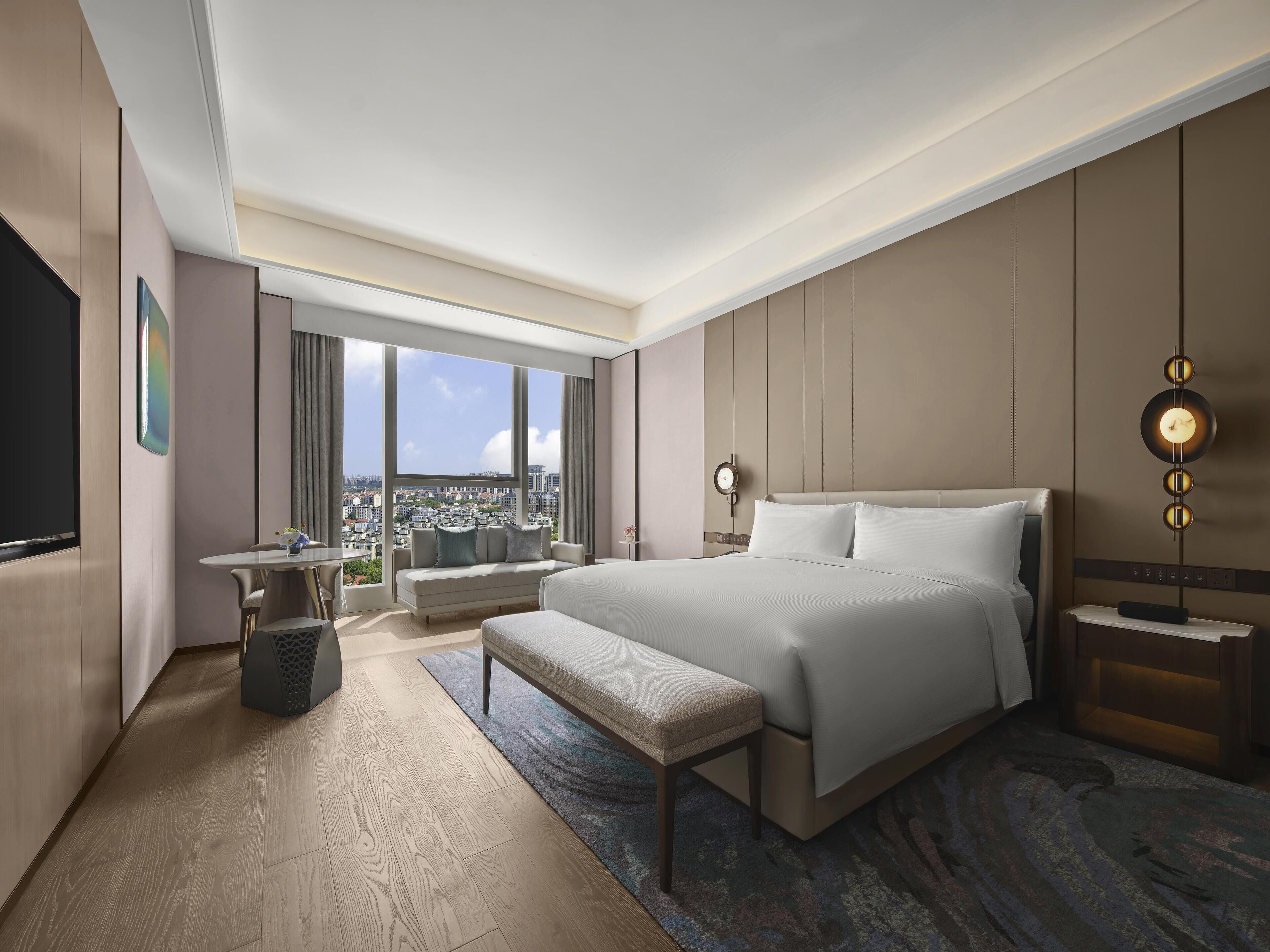 Quarto de hotel moderno com uma cama grande, área de estar, televisão e vista para a cidade.