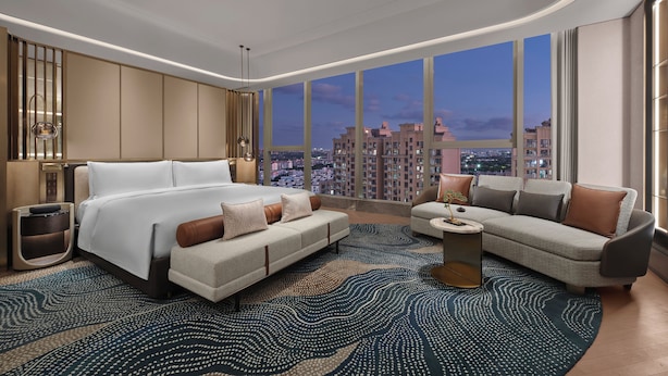 A modern hotel room with a large bed, a sofa, a coffee table, and a view of the cityscape.