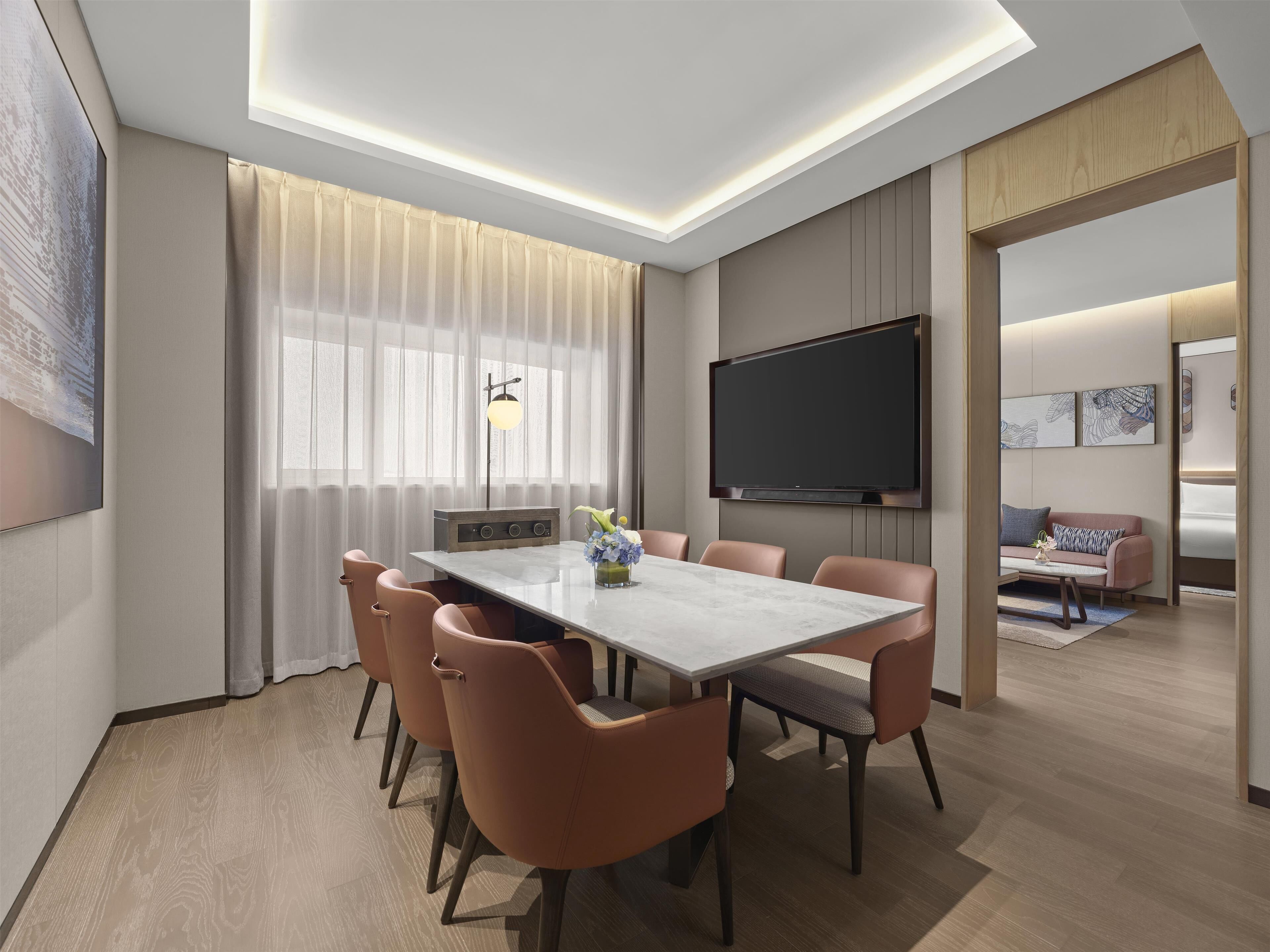 A modern conference room with a large table, six chairs, a flat-screen TV, and a large window with sheer curtains.
