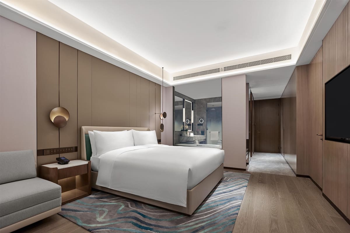 Une chambre d’hôtel moderne dotée d’un grand lit, d’une table de chevet avec un téléphone, d’un coin salon, d’une salle de bain avec douche et d’une télévision.