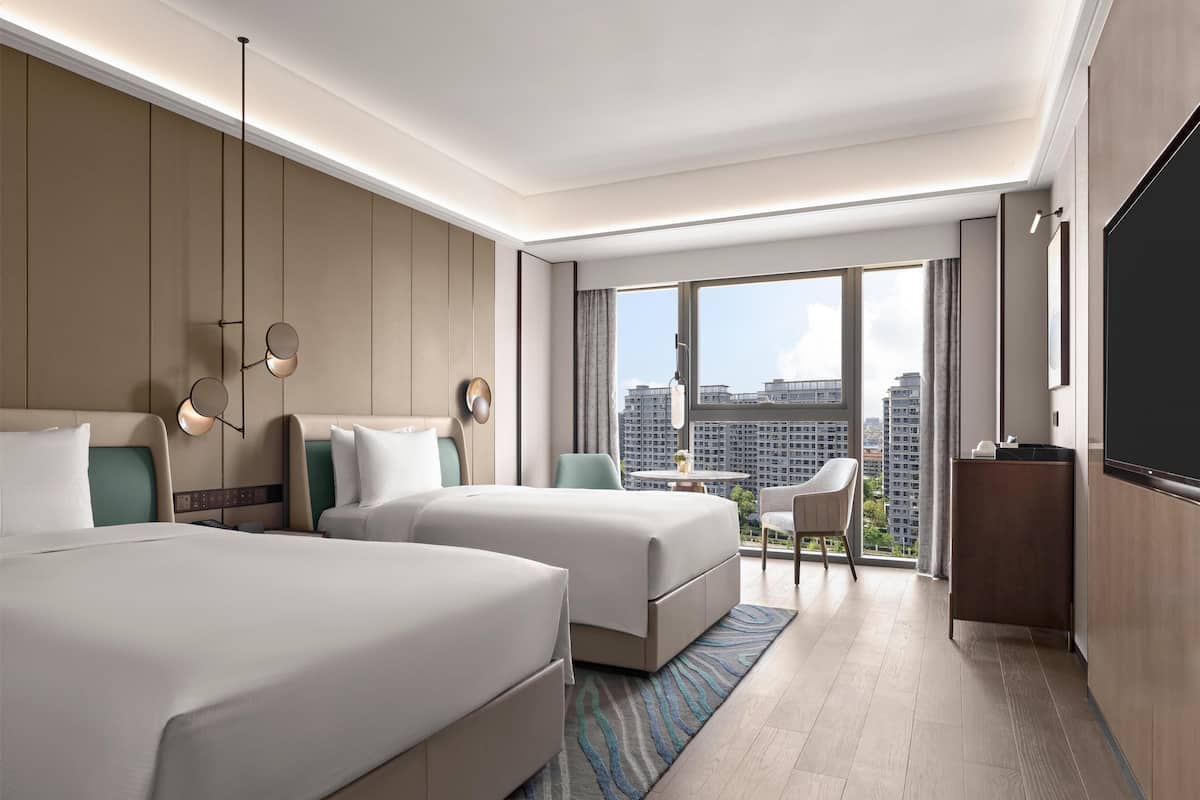 Une chambre d’hôtel moderne avec deux lits, une petite table, une chaise, un téléviseur et de grandes fenêtres donnant sur la ville.