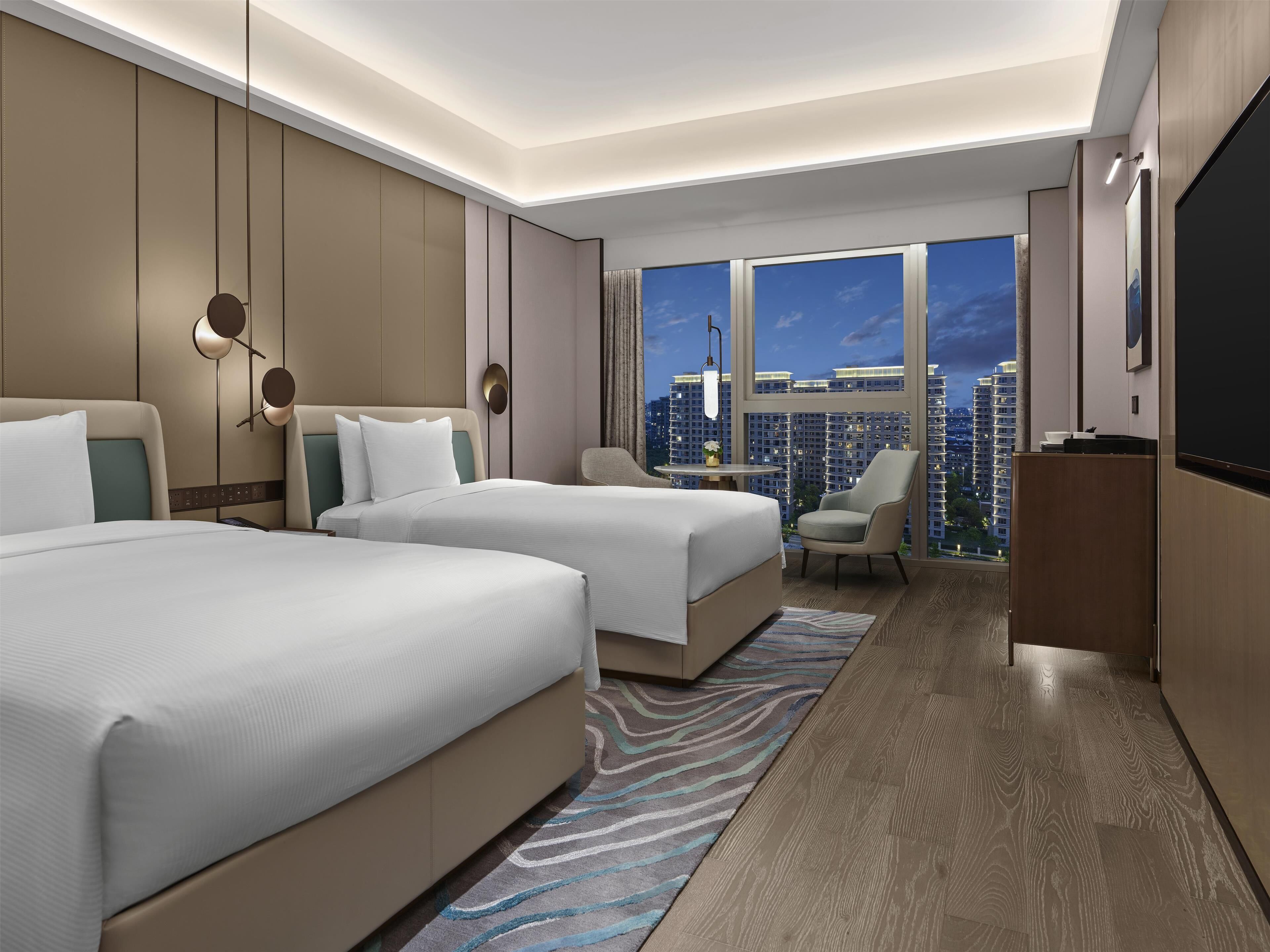 Una habitación de hotel moderna con dos camas, un escritorio, una silla y un ventanal con vistas a la ciudad.