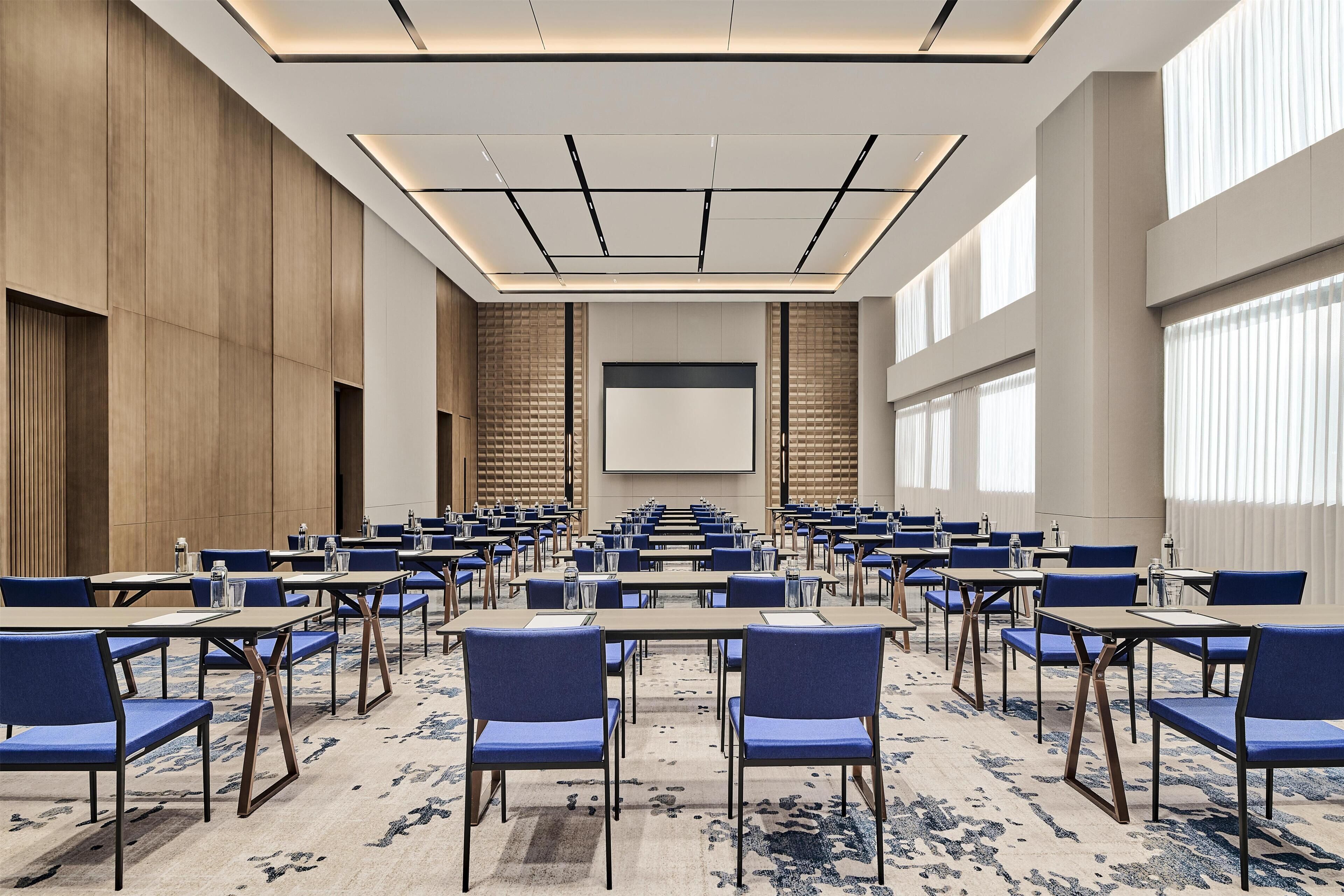 Une salle de conférence avec des rangées de tables et de chaises, un grand écran et une moquette à motifs.