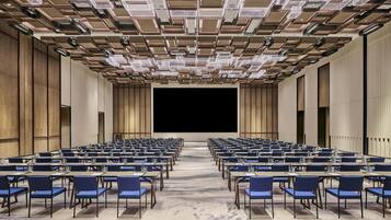 A conference room with rows of tables and chairs, a large screen at the front, and a patterned ceiling.