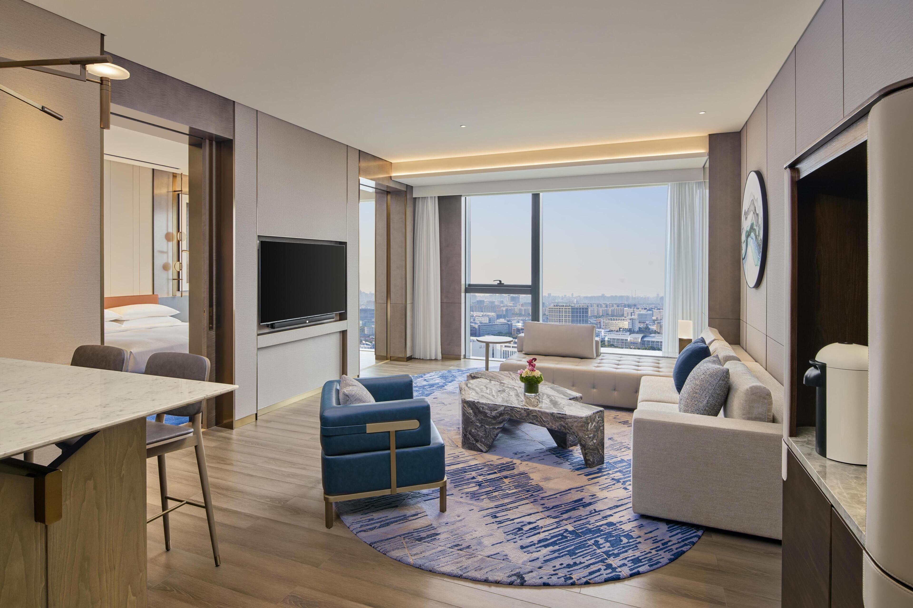 A modern hotel room with a large window, a flat-screen TV, a sofa, a coffee table, and a dining area.