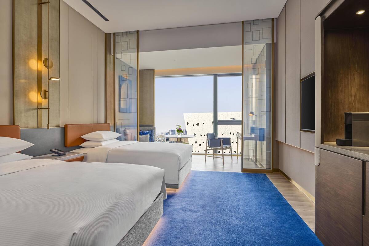 Ein Hotelzimmer mit zwei Betten, einem Fernseher, einem Balkon mit Blick auf die Stadt und einem blauen Teppich.