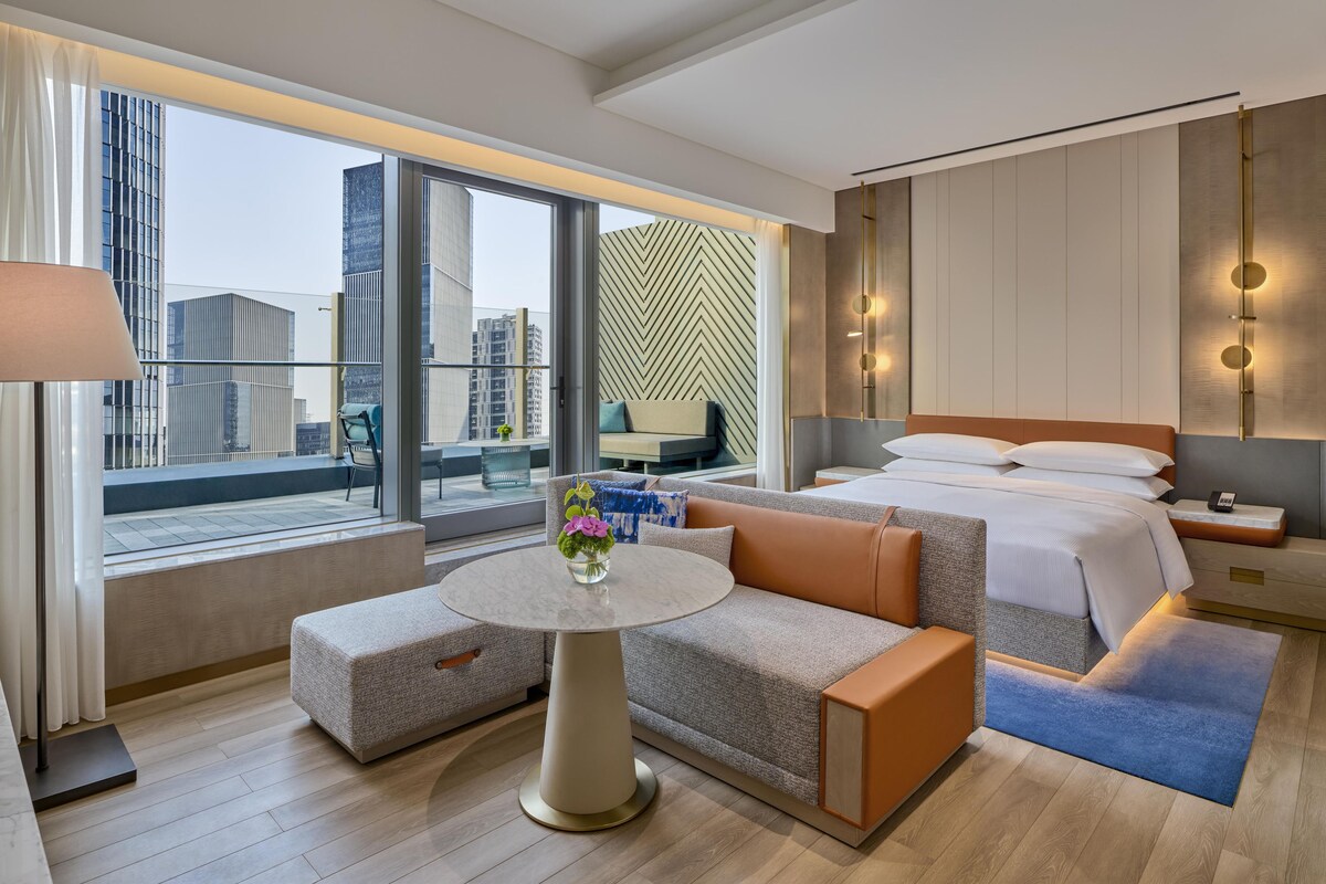 Une chambre d’hôtel avec un lit, un canapé, une petite table et une vue sur le paysage urbain grâce à de grandes fenêtres.