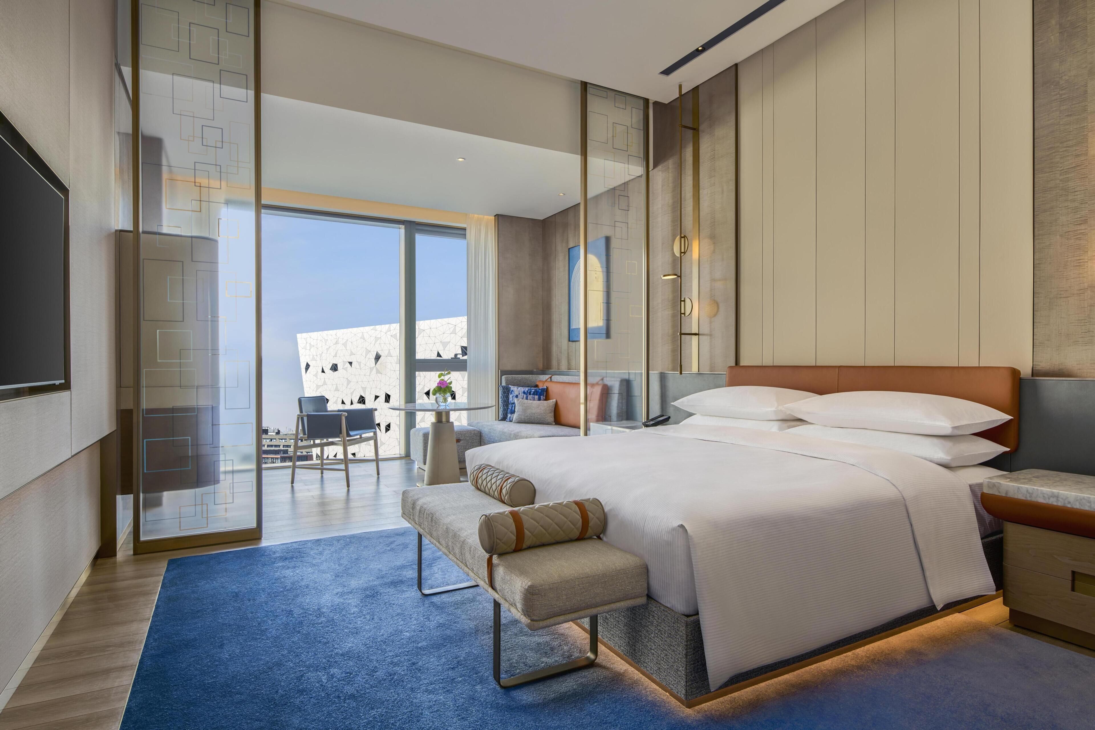 A modern hotel room with a large bed, a seating area, a desk, and a view of the city.