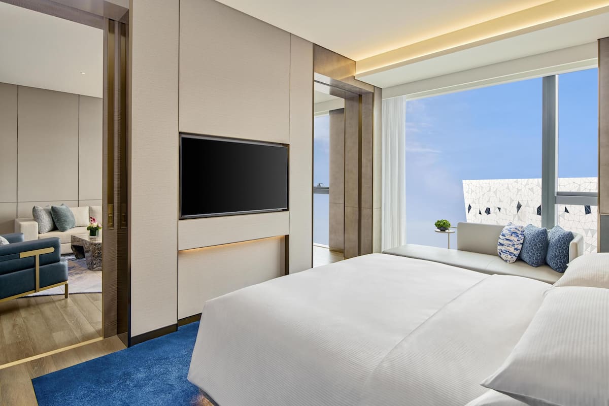 Ein modernes Hotelzimmer mit einem großen Bett, einem an der Wand befestigten Fernseher, einer Sitzecke mit Sofa und Sesseln sowie einem Balkon mit Blick.