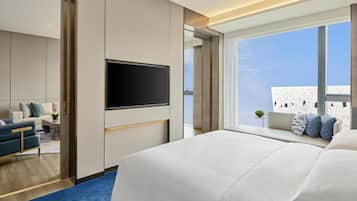 A modern hotel room with a large bed, a TV mounted on the wall, a seating area with a sofa and armchairs, and a balcony with a view.