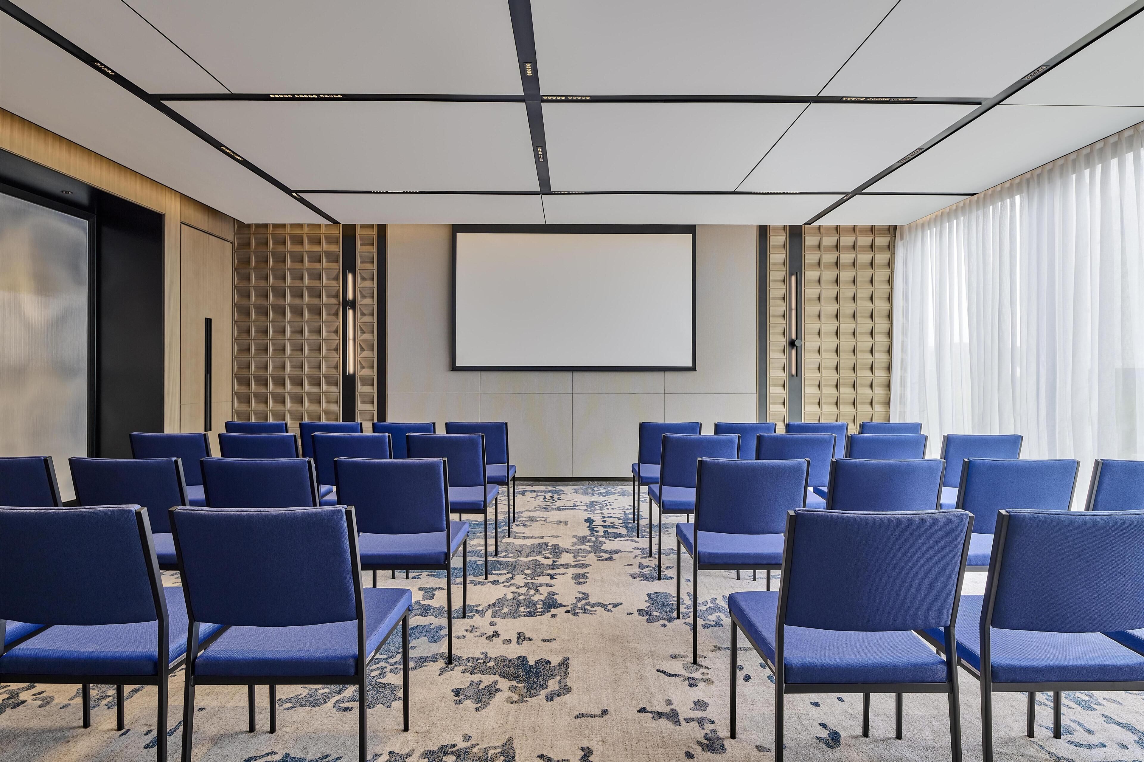 A conference room with rows of blue chairs, a large screen, and a patterned carpet.