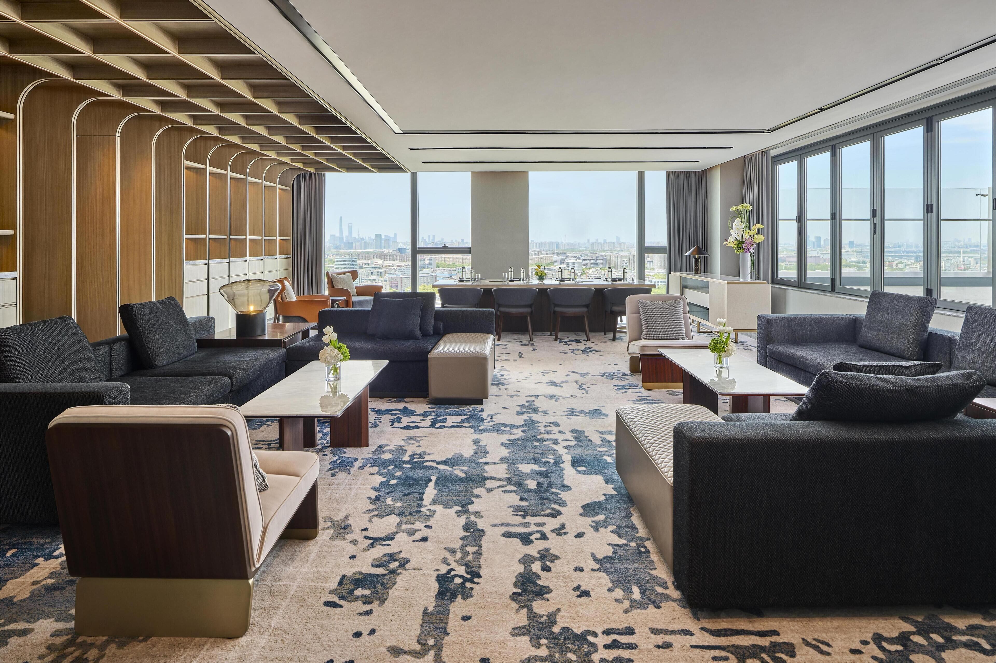 A modern hotel lobby with a large seating area, a central table, and large windows offering a city view.