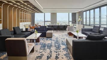 A modern hotel lobby with a large seating area, a central table, and large windows offering a city view.
