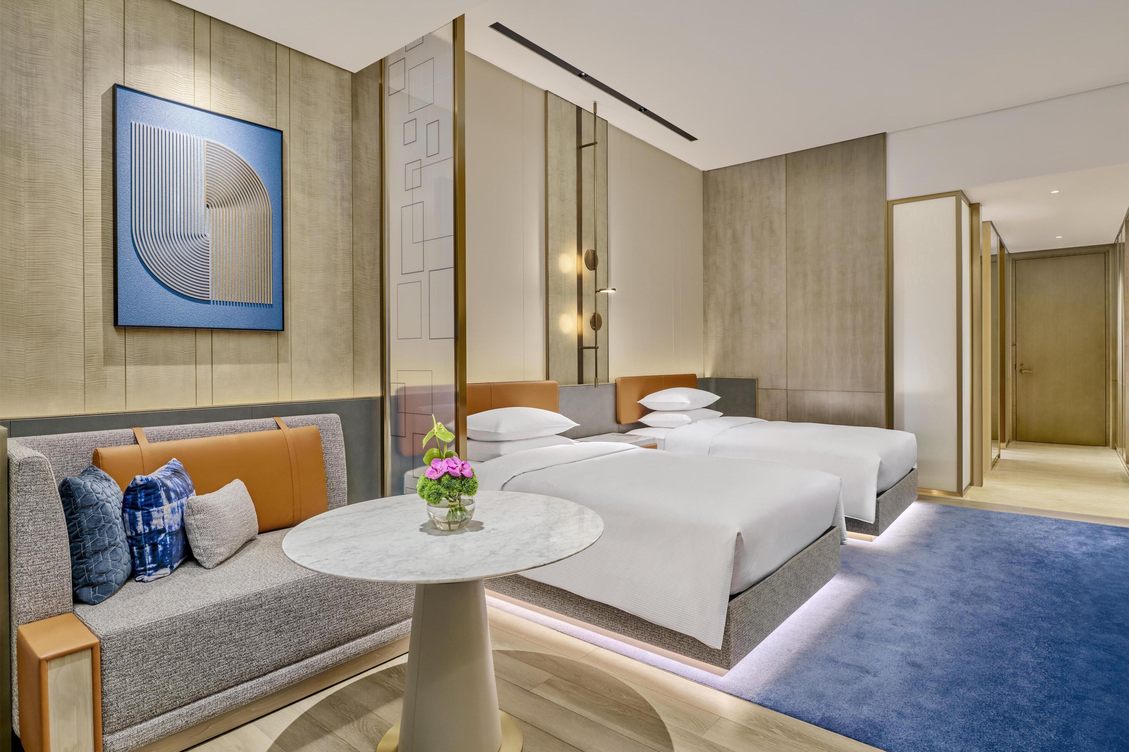A hotel room with a sofa, a round table with a vase, two beds, and a large abstract painting on the wall.