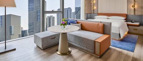 A modern hotel room with a large window, a round table, a sofa, a bed, and a nightstand.
