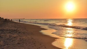 Una playa con un muelle que se adentra en el agua, olas suaves lamiendo la orilla y el sol poniéndose en el horizonte.