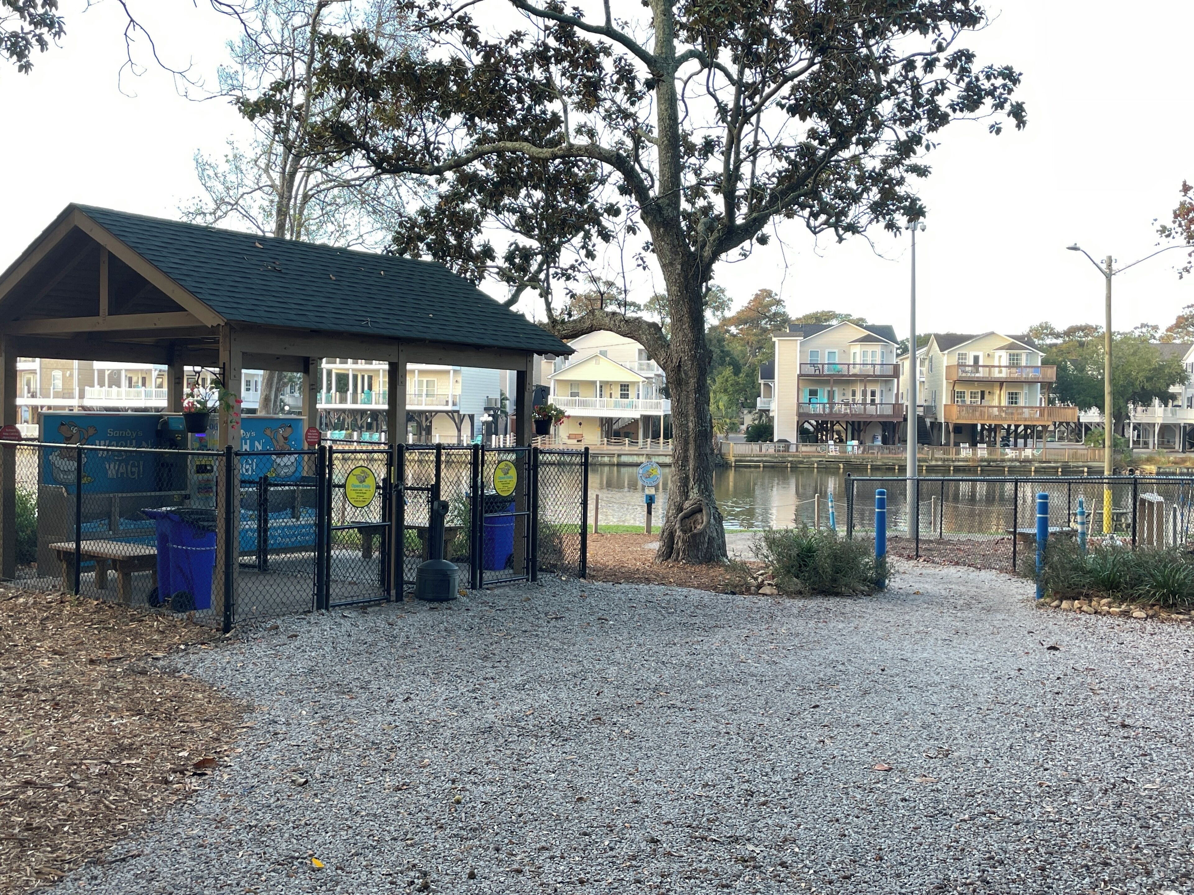 A covered seating area with a gravel path, a fenced playground, and a body of water with buildings in the background.