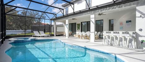 A swimming pool with a glass enclosure, lounge chairs, and a covered patio area.